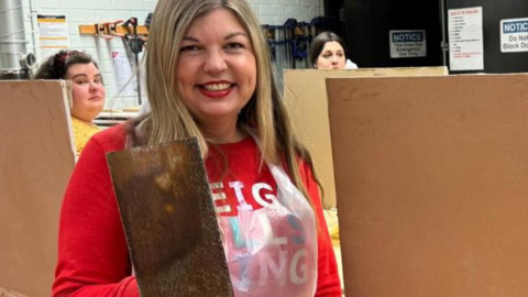 Emma Meese in a red jumper and plastic apron, holding construction equipment 