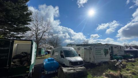 Lots of vans and trailers parked together on a sunny day with large buildings in the background.