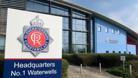 The exterior entrance of the Gloucestershire Constabulary HQ in Waterwells. IT is a large blue building with a sloped roof and lots of windows. There is a Gloucestershire Police sign outside, with a manicured hedge and a paved pathway in the background. 