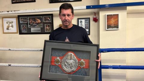 A man in a black t-shirt is holding a kickboxing title belt, encased inside a frame. He is standing inside a boxing ring.