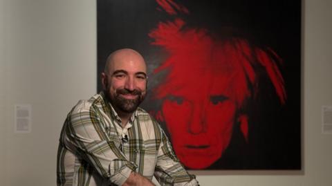 A man smiling in a gallery in front of a black and white self portrait by Andy Warhol