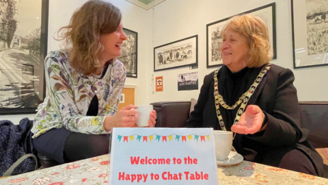 A woman with shoulder-length mousy brown hair wearing a blue skirt and a white blouse with a flowery pattern sits at a table holding a mug. She is laughing and talking to another woman with slightly shorter blonde hair with a mug on the table in front of her. She is wearing a black outfit and round her neck is a gold mayoral chain. A sign on the table reads: "Welcome to the Happy to Chat Table".