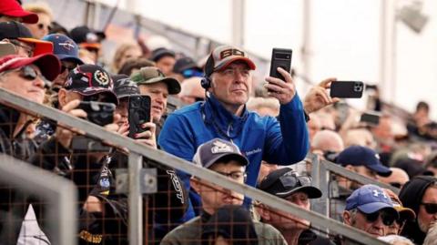 Race fans sitting on the grandstand holding up mobile phones to film the bikes.
