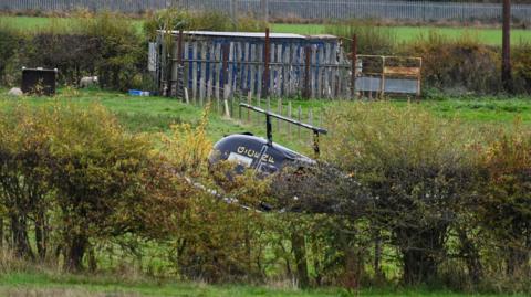 A black helicopter on its side in a field, partly concealed behind some bushes.