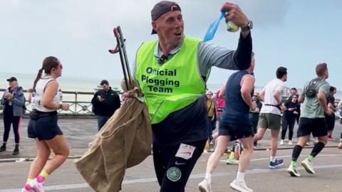 Darren Evans wearing a green bib saying 'official plogging team' whilst running. He looks to his left and holds up litter.
