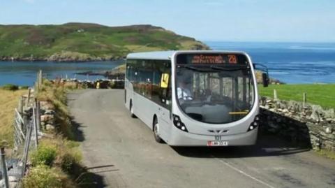 The number 28 Bus Vannin bus travelling towards Port St Mary from the Sound. The Calf of Man can be seen in the background with a channel of water between the two land masses. There are stone walls and farmers' fencing at either side of the road the silver single-decker bus is travelling on. The bus has the number 28 and via Cregneash and Port St Mary written on the front on an LED screen above the windscreen, 