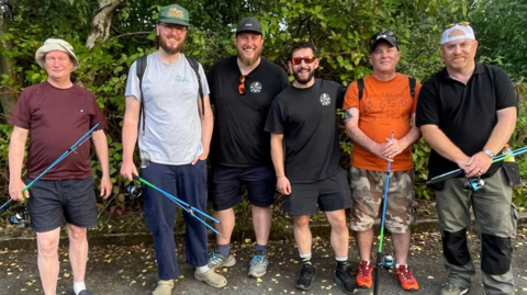 A group of six men stand in front of trees holding fishing rods. 