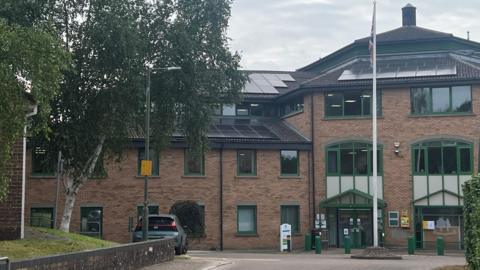 The Forest of Dean District Council building in Coleford. It is a red-brick building with a black roof and green window frames. The sky behind it is grey and there are green trees to the left.