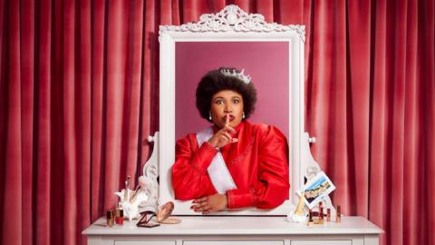 Anne Odeke wearing a crown and a red dress. 
She is standing by a vanity table and has emerged herself in the mirror and has her index finger on her lips.