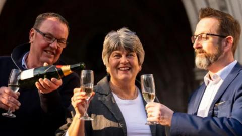 A woman stands in the middle of two men. She is wearing a dark jacket and white tee-shirt, one of the men wears a blue suit, the other is wearing a dark jumper. They are pouring a bottle of champagne. They are standing in front of an ornate, carved stone arch