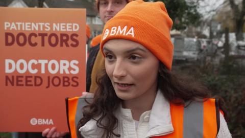 Dr Heather Gunn has long, curly brown hair under her orange BMA beanie hat. She is wearing an orange fluorescent jacket and standing on the picket line, next to a sign that reads "Patients need doctors. Doctors need jobs."