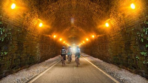 A group of cyclists pictured riding away from the camera through a large tunnel. The tunnel has a tarmac path and is lit by lights along the walls of the tunnel.