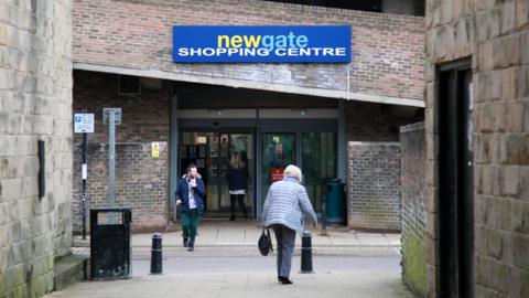 An entrance to Newgate Shopping Centre with a woman holding a handbag heading towards the doors and a man leaving. A large blue sign above the entrance carried the centre's name in yellow, light blue and white letters. It looks like a side entrance with two automatic doors and a rising sloped brick wall (like the outside of a car park ramp) above.