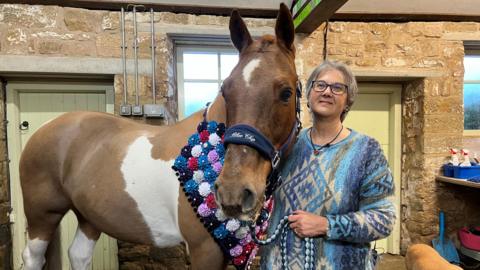A big horse with a brown coat with patches of white stood next to a woman in a blue patterned jumper. They are stood inside some stables.