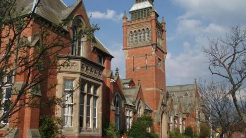 A redbrick school building with large bay windows. A clock tower rises above the building, which is set behind some trees.