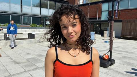 Chloe Ing, 21, illustration student, looks straight at the camera. She has long dark curly hair and is wearing an orange vest top