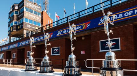 Large silver Classic TT trophies line up in front of the grandstand, which has TT  branding running along it.