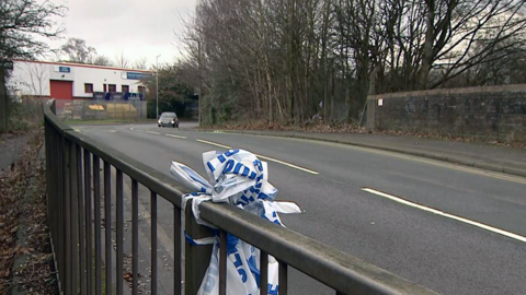 Police tape tied on to a metal bridge on a curved industrial road lined with trees and bushes. There is white warehouse in the background.
