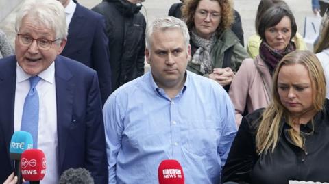 Hugh Harper, whose daughter, Leona, was killed in the blast is in the middle of the picture. He is wearing a blue shirt and has fair hair. He is looking into the distance. He is with picured with other bereaved families. ONe of them, a woman at the front, is holding up a picture of her loved one. Solicitor Damian Tanswey (far left) is wearing a dark suit, blue tie, white shirt and has glasses. There are microphones in front of the group as they speak to the media.