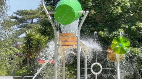 A water park with water pouring out of a big green bucket. A variety of trees are visible in the background, as well as green and orange plastic wheels that appear to be squirting water around.