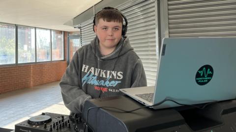 Albie is wearing headphones and a grey sweatshirt with 'Hawkins Hitmaker' written on the front. He is standing behind a silver laptop and DJ decks and in front of a window and a closed shop front. 