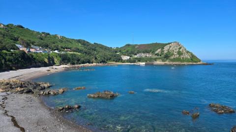 A curved beach with rocks in sea, steep coastline behind with trees and houses, under a clear blue sky