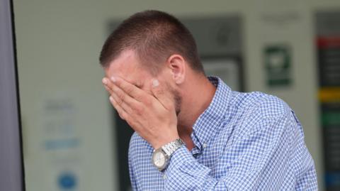 Shaun Thompson, who is wearing a blue and white checkered shirt, covering his face as he leaves court. He has short brown hair.