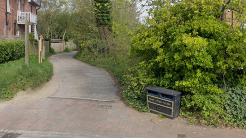 A photo taken from a road looking down a narrow lane with trees on the right. On the left is a brown brick house. There is a large black litter bin on the right of the entrance.