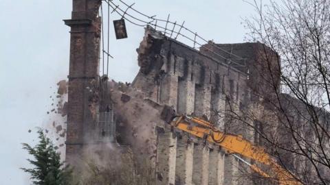 The wall of a historic mill building is being pulled down by a yellow mechanical device. There are bricks and debris falling as the wall begins to collapse.