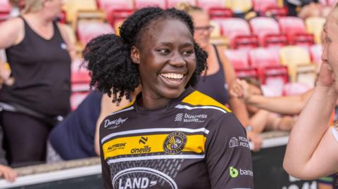 Peace Lekuru smiles at a teammate on the rugby pitch. She wears a York Valkyrie rugby shirt, which is black with a yellow stripe across the chest, with white sponsor logos across the front and sleeves. Spectators can be seen behind her in the stands.