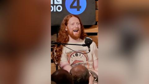 A man with long ginger hair laughs into a microphone.