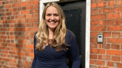 A woman with long blonde hair and wearing a navy top is smiling widely. She is standing in front of a red brick wall and a black door with a white frame