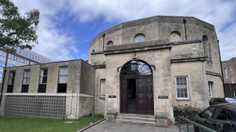 An old court building with a big arched door, grey stone walls and lots of windows. Behind it is a modern building and a tree. 