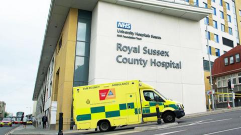 A yellow ambulance enters an access road on a slope, alongside a modern building with the words "Royal Sussex County Hospital" written large on a cream-coloured stone wall.