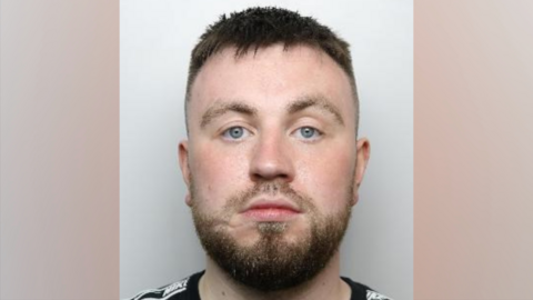 A police mugshot of Ethan Hallows. He has short, brown hair, closely shave at the side of his head. He has a dark, brown beard and facial hair.