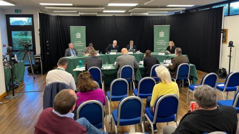 A table with a green tablecloth with five people sitting behind it and banners which say Scrutiny Management Committee behind it. In front is another table with a similar tablecloth, with four people sat behind it. Behind them are rows of blue cushioned chairs with some taken up by members of the public and politicians. 