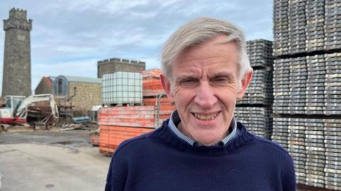 Smiling man in navy jumper, known as a "Guernsey Knit", stands in a construction yard with scaffolding, machinery, and stone tower behind him under an overcast sky.
