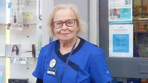Sadie Jefferson has long blonde hair. She is wearing red-rimmed glasses and is wearing her blue pharmacy uniform. She has a name badge and lanyard and is standing outside the Pharmacy doors. 