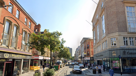 A Google Streetview showing Granby Street heading towards Leicester Railway Station. There are cycle lanes, cars and pedestrians shown with shops either side of the street.