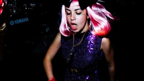 Charli XCX in a purple glitter dress, a pink wig and a golden chain, in the midst of singing.