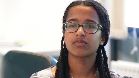 Alyssia, 17, is wearing glasses and has long braids. She is sitting at a table taking part in a discussion 