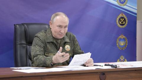 Vladimir Putin wears army fatigues speaking holding newspaper in his hand