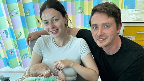 A woman in a white top holds a small baby wearing medical equipment. She is sat next to a man in a black top. A hospital curtain is visible in the background.