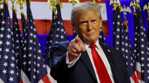 Donald Trump smiles as he points on stage in front of a row of American flags