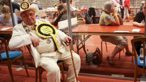 Alan Hope has a white stetson, suri, waistcoat and shirt and is seen here seated at the Wakefield by-election count in 2022.