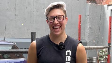 Laura Hollywood stands outdoors in front of a grey wall, wearing a sleeveless black shirt with white vertical text on the front.