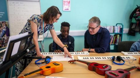 The prime minister sits at a table with guitars and headphones on it. A young boy sits next to him and a woman stands on the boy's other side, pointing at a sheet of music.