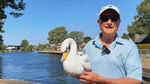 A man with a blue shirt, baseball cap and sunglasses. He is holding a toy swan.