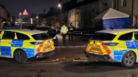 An evening scene of two police cars, a handful of police officers and a blue gazebo-style forensics tent on a street of terraced housing.