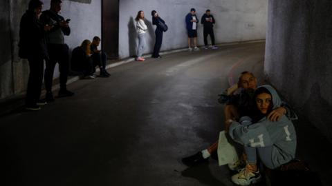 People stand and sit in pairs as they shelter in an underground concrete space 